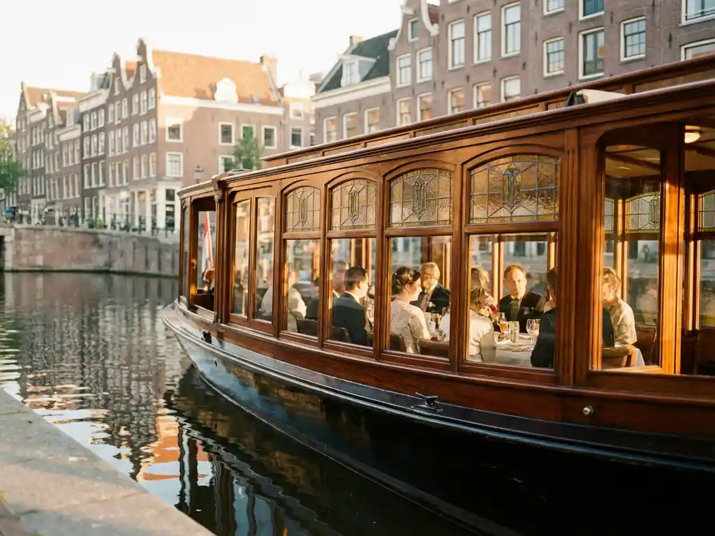 Vintage 1910 Dutch salon tjalk with polished iroko hull gliding through an Amsterdam canal at golden hour, guests dining inside glazed salon windows.