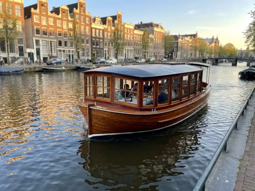 Dutch salon tjalk canal boat with polished iroko wood hull gliding past Amsterdam's gabled houses at golden hour, guests dining inside.