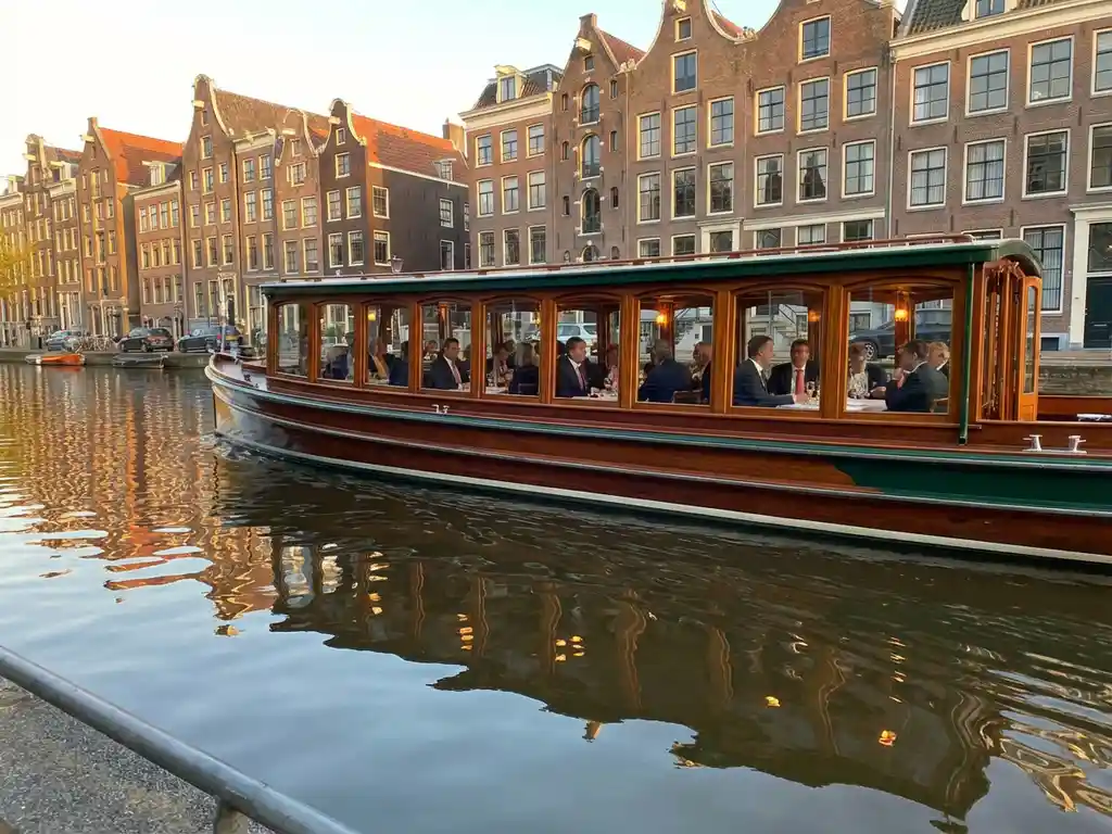 1910 Dutch salon tjalk with polished iroko hull cruising an Amsterdam canal at golden hour, historic gabled houses reflected in calm water.