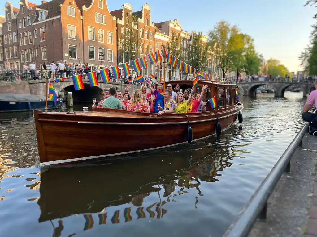 Luxury Dutch salon boat cruising Amsterdam canal during Pride, rainbow flags lining historic gabled banks, guests celebrating on polished iroko wood deck.