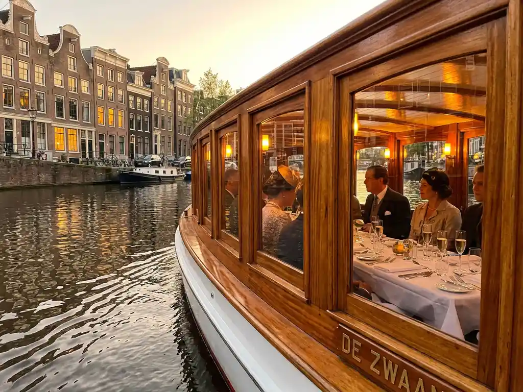 Classic 1910 Dutch salonboot with polished iroko wood gliding Amsterdam canals at golden hour, guests dining inside with champagne and catering platters.