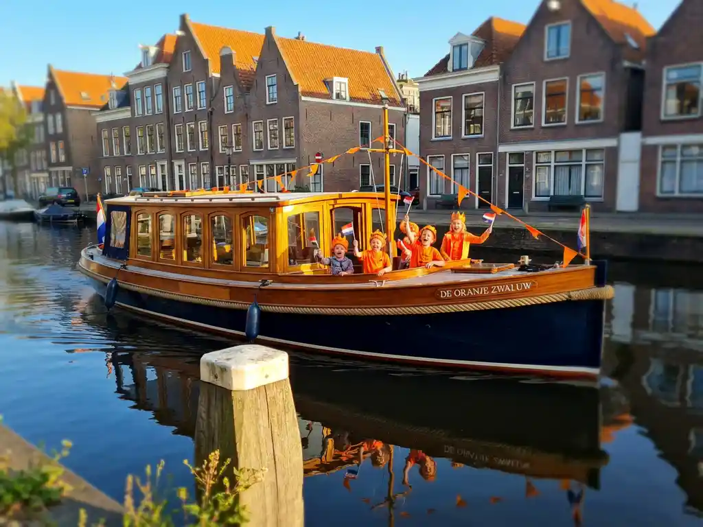 Dutch salon tjalk sailing Amsterdam canals on King's Day, children in orange outfits laughing on iroko wood deck, gabled houses reflecting in calm water.