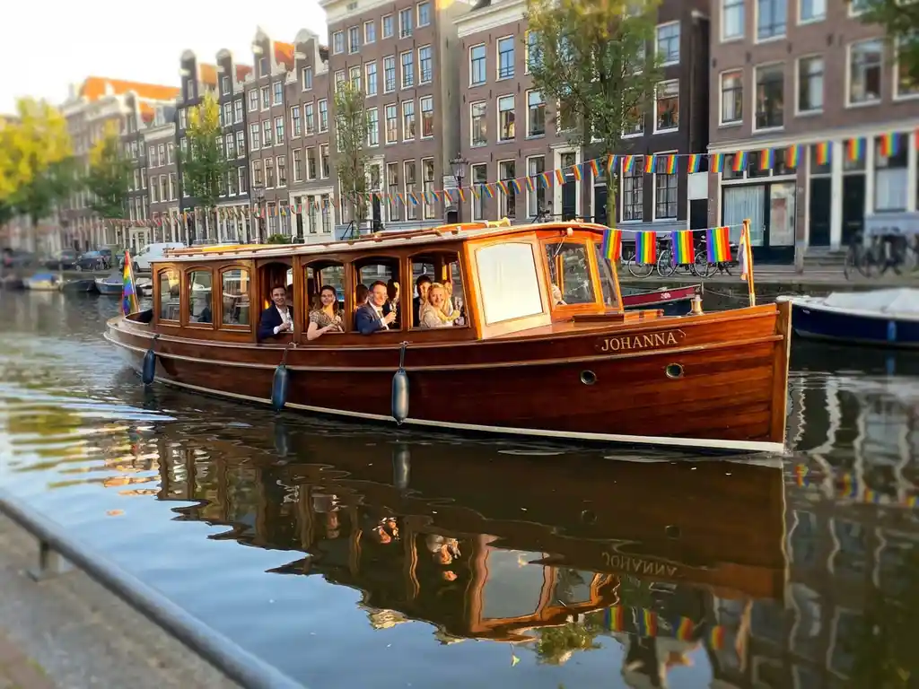 Classic 1910 Dutch salon tjalk with polished iroko wood gliding through rainbow-flag-lined Amsterdam canals during Pride, golden afternoon light.