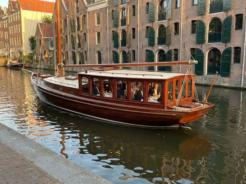 1910 Dutch salon tjalk gliding through Amsterdam canals at golden hour, polished iroko hull reflecting in calm water beside historic gabled facades.