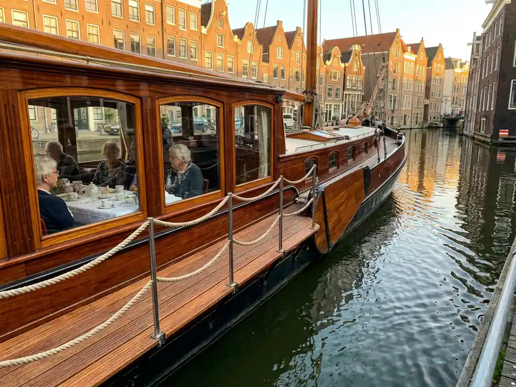 Dutch salon tjalk from 1910 cruising Amsterdam canals at golden hour, polished iroko hull reflecting historic gabled houses, elderly guests dining inside glazed salon windows.