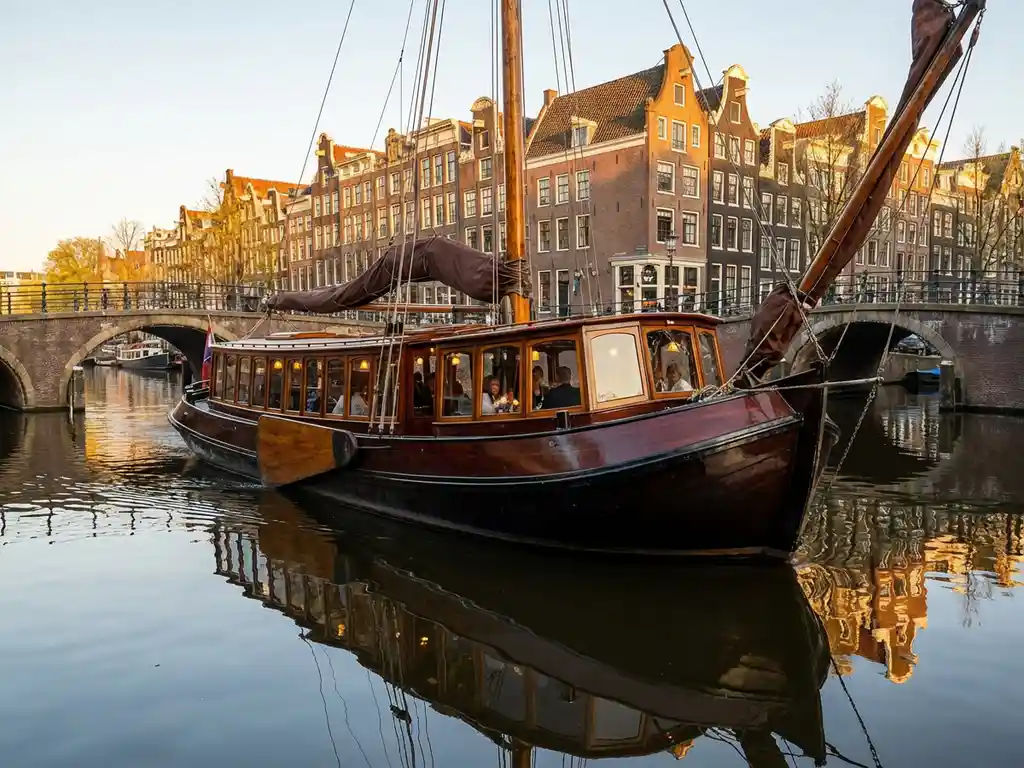 1910 Dutch salon tjalk with polished iroko wood hull cruising Amsterdam's historic canals at golden hour, guests dining inside glazed salon windows.
