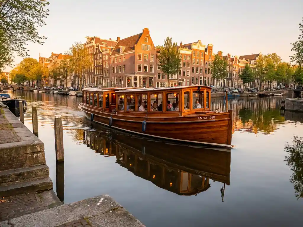 Classic 1910 Dutch salon tjalk with polished iroko hull cruising Amsterdam canal at golden hour, guests dining inside, historic gabled facades reflected in calm water.