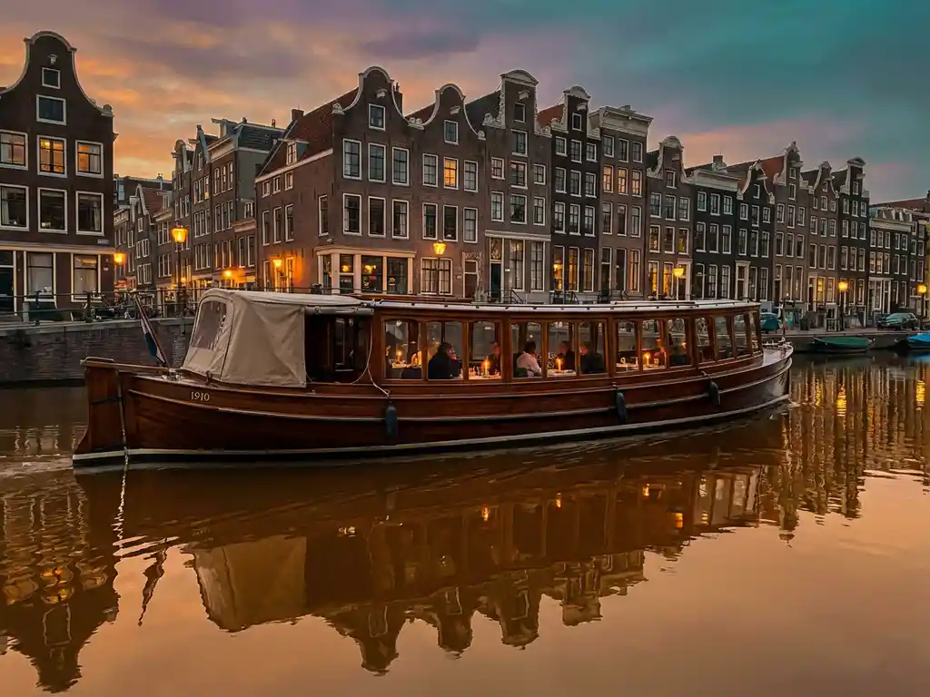 1910 Dutch salon tjalk with polished iroko hull cruising Amsterdam canals at golden hour, guests dining inside as gabled houses reflect in amber water.