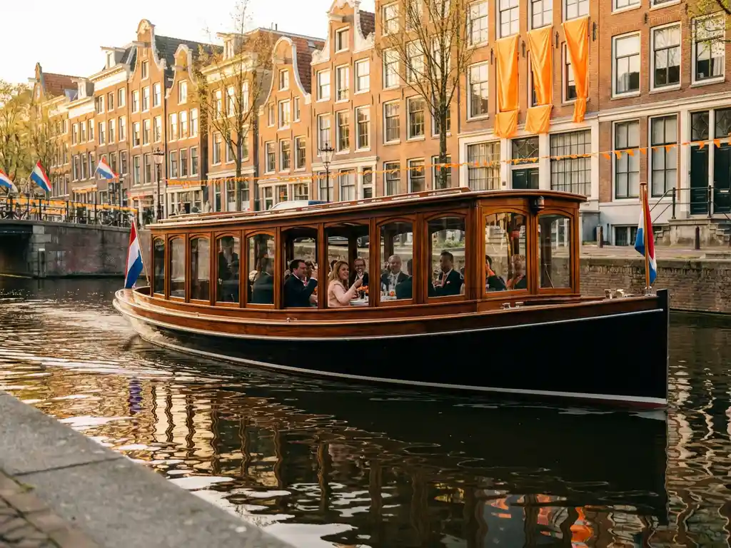 1910 Dutch salon tjalk with polished iroko hull cruising Amsterdam canals on King's Day, guests dining inside glazed windows, orange-draped gabled houses reflected in water.