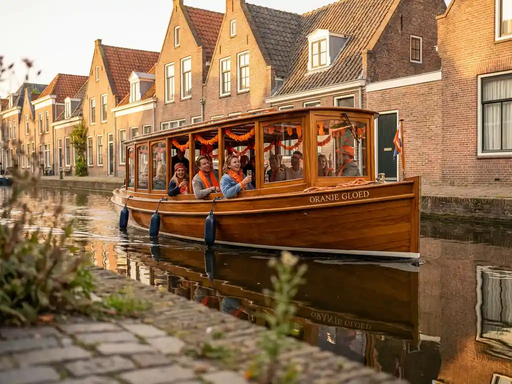 Friends celebrating King's Day aboard a Dutch salon tjalk with orange garlands, gliding past Amsterdam's historic gabled canal houses.