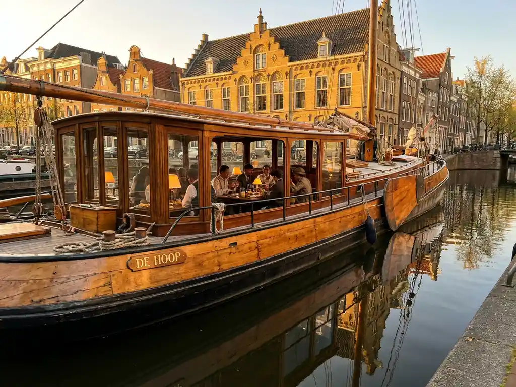 Dutch salon tjalk from 1910 gliding through Amsterdam canals at golden hour, polished iroko hull gleaming beside historic gabled facades.