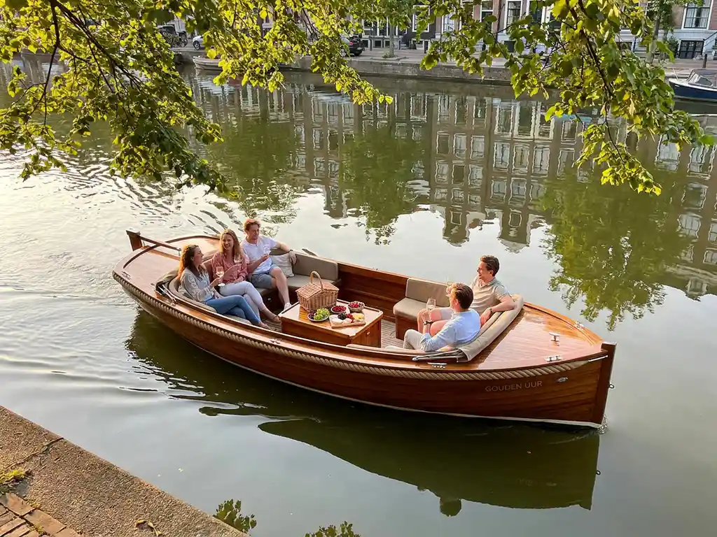 Friends enjoying wine and a picnic aboard an electric sloop on a narrow Amsterdam canal, historic Dutch gabled buildings reflected in calm green water at golden hour.