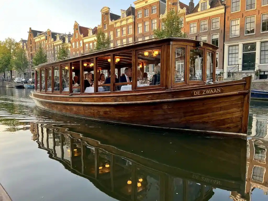 1910 Dutch salon tjalk gliding through Amsterdam canals at golden hour, polished iroko hull reflecting historic gabled houses in calm water.