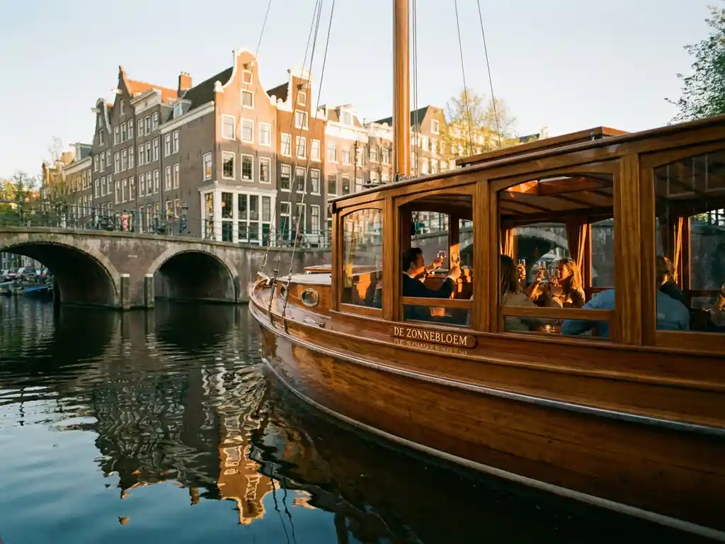1910 Dutch salon tjalk with polished iroko hull gliding through Amsterdam canals at golden hour, historic gabled facades reflected in glassy water.