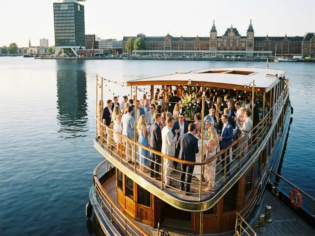 Luxury event boat cruising Amsterdam's IJ waterway at golden hour, elegantly dressed guests on open deck, A'DAM Tower and Central Station reflected in calm water.