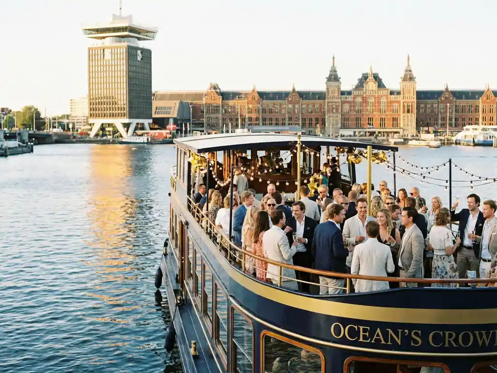 Luxury party boat on Amsterdam's IJ waterway at golden hour, guests on upper deck with string lights, A'DAM Tower and Central Station in background.