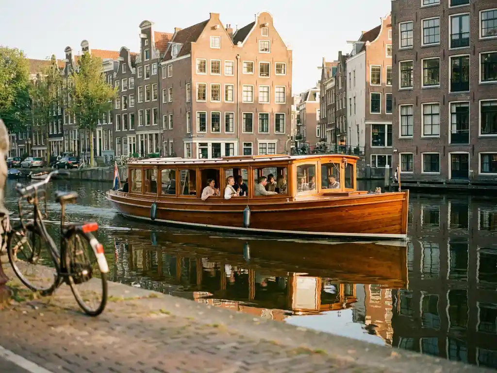 1910 Dutch salon tjalk with polished iroko wood hull cruising Amsterdam canals past historic gabled facades in warm afternoon light.