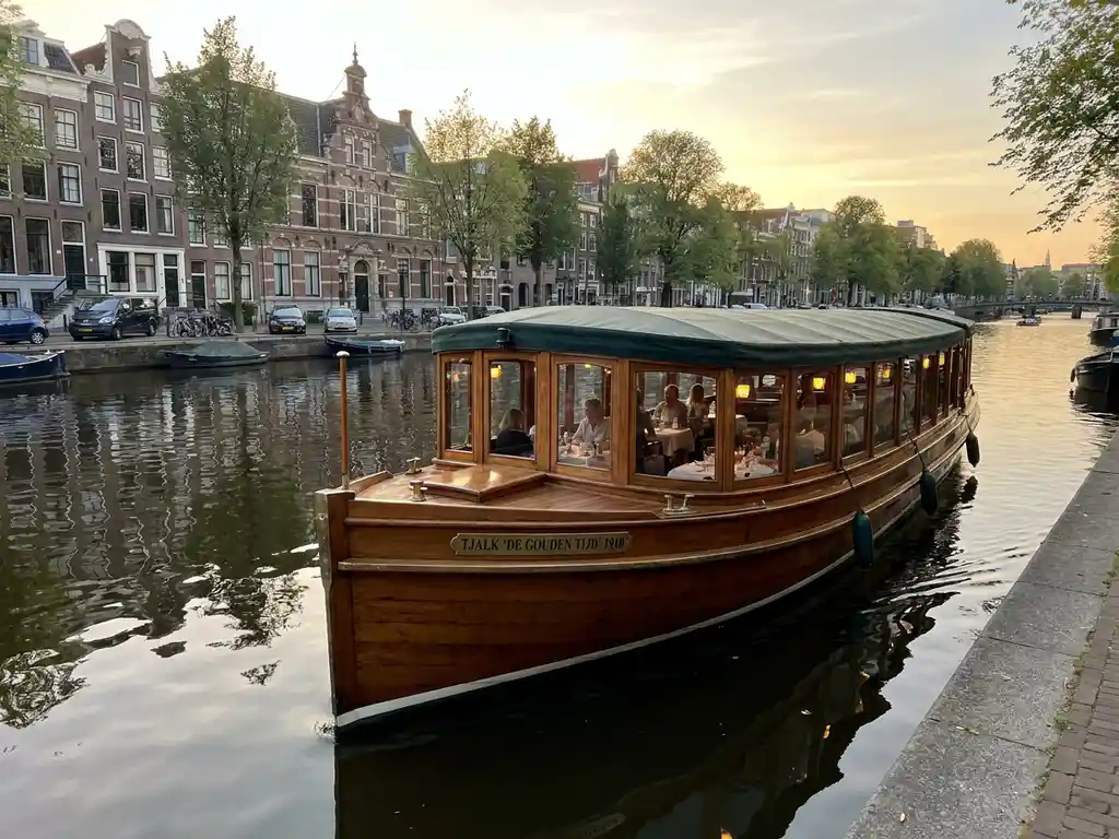1910 Dutch salon tjalk with polished iroko hull gliding through Amsterdam canals at golden hour, guests dining inside glazed windows, historic gabled facades reflected in calm water.