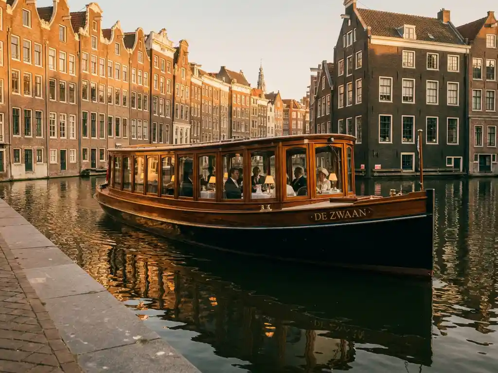 1910 Dutch salon tjalk gliding through an Amsterdam canal at golden hour, polished iroko hull reflecting in glassy water, historic gabled facades lining the banks.